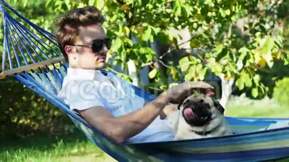 戴墨镜的冷静男子和一只小狗在吊床上休息在花园里的日落时放松视频的预览图