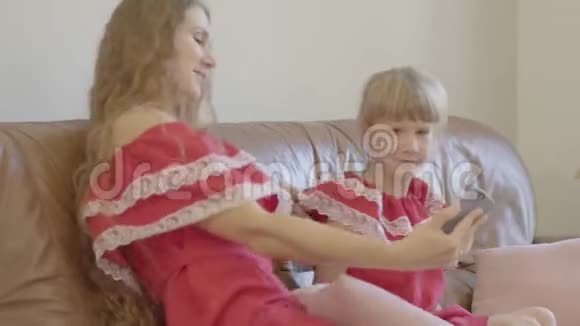 白人母亲和小女儿穿着红色连衣裙坐在沙发上在电话里自拍开朗的女人视频的预览图