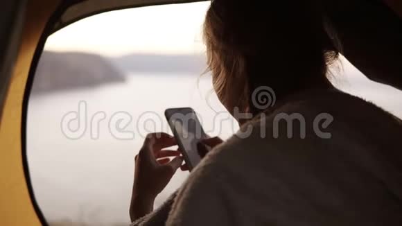 野营的女人坐在帐篷里看着用她的智能手机拍的大自然的照片露营女孩在度假中放松视频的预览图