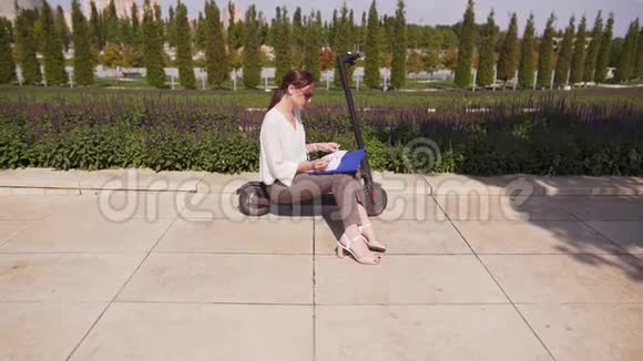 在一个阳光明媚的夏天年轻漂亮的女人坐在公园里的电动踏板车上学生创建记事本视频的预览图