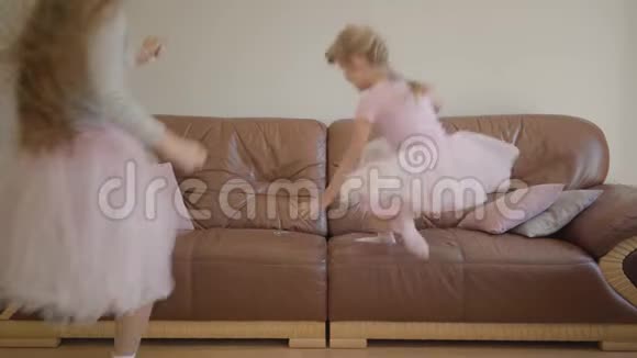 快乐的白种人妈妈和女儿穿着同样的粉红色连衣裙跳上教练用遥控器转动电视视频的预览图