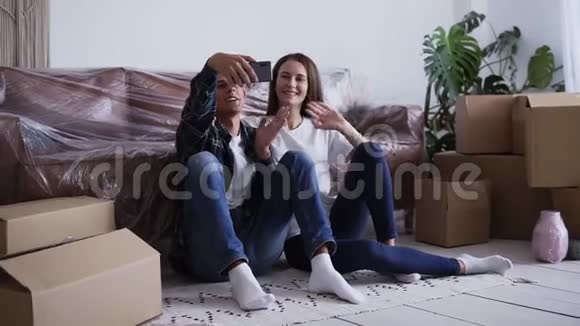 年轻快乐的白种人夫妇坐在他们的新房子的地板上自拍着一堆未包装的纸板箱视频的预览图