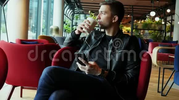 迷人的男人在现代时尚的咖啡馆里喝咖啡用手机浏览互联网视频的预览图