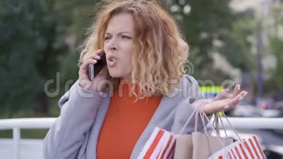 不满意的红发女人拿着购物袋用手机说话购物狂站在那里购物视频的预览图