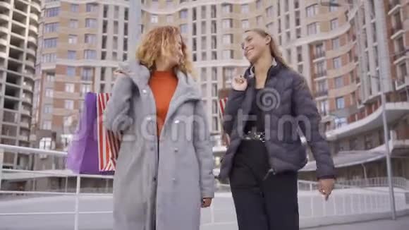两个快乐美丽的女孩拿着购物袋走在街上说话微笑无忧无虑的生活方式Shopaholism视频的预览图