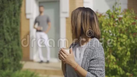 可爱的白种人女人站在前台喝茶她的丈夫从后面走过来亲吻她视频的预览图