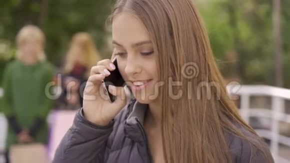 年轻迷人的白种人女孩在电话旁交谈她的朋友在后台等她休闲在一起视频的预览图