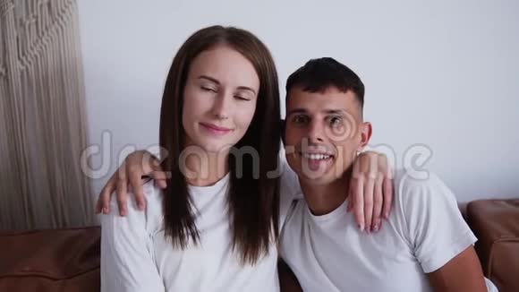 这对相爱的年轻夫妇拥抱的肖像对着白色背景墙上的相机微笑肖像在里面视频的预览图