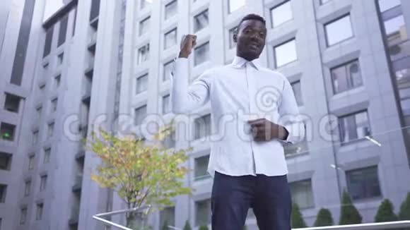 快乐快乐的非洲裔美国人穿着白色衬衫站在城市街道上举起拳头打胜利的手势视频的预览图