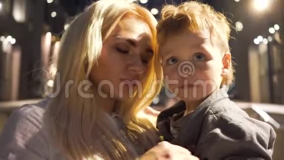 一个漂亮的金发女人穿着白色衬衫双手抱着她的小男孩母亲和儿子晚上谈话视频的预览图
