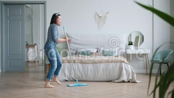 年轻的黑发女人在卧室里拖着拖把唱歌在家里洗着地板跳舞视频的预览图