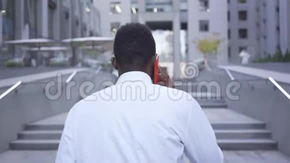 美国黑人男子穿着白色衬衫走上楼梯用红色手机说话男性商人或视频的预览图