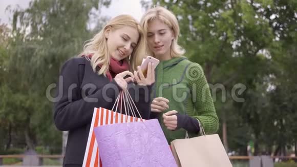 两个漂亮的金发女朋友站在街上拿着购物袋看着手机两位时尚女性视频的预览图