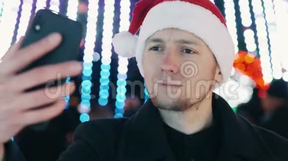 开朗的男人自拍并在手机上建立有趣的面孔穿着圣诞帽圣诞树视频的预览图