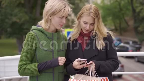 两个漂亮的金发女朋友站在街上用购物袋买东西付钱的女人视频的预览图