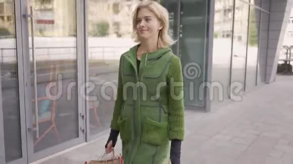 自信的年轻金发女人穿着绿色夹克走在街上城市居民的休闲时尚概念视频的预览图