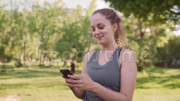 阳光明媚的清晨在公园里一位高加索健康的年轻女子一边微笑一边用手机发短信视频的预览图