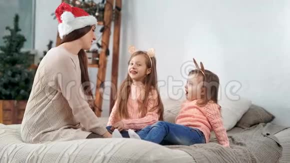 年轻的妈妈戴着圣诞老人的帽子两个可爱的女儿戴着有趣的节日耳朵玩视频的预览图