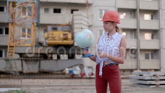 戴着建筑头盔的可爱漂亮的年轻女子手里拿着地球仪把它扭曲指向乡村或城市视频的预览图