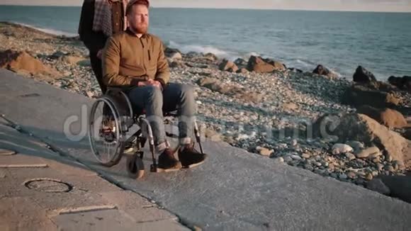年轻女子正在护理生病的残疾男子在海边滚动婴儿车视频的预览图
