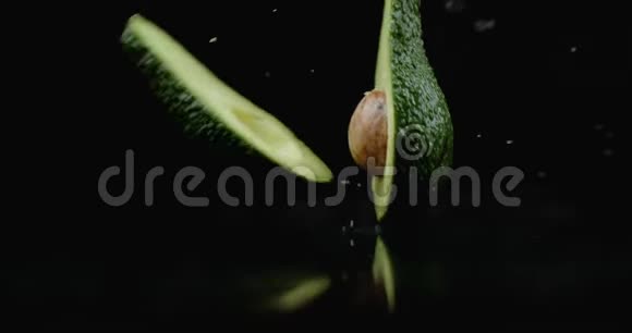 一个成熟的切割绿色鳄梨落在玻璃上水在黑暗的背景上缓慢地飞溅视频的预览图
