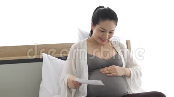 孕妇在卧室的家中在床上用手轻轻触摸腹部同时看乳房X光照片人们的生活方式视频的预览图