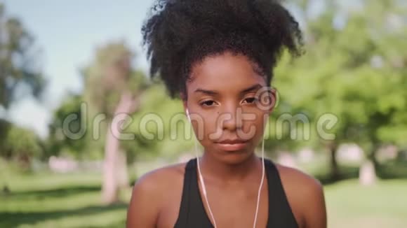 年轻健康的非裔美国妇女在阳光明媚的公园里微笑着欣赏耳机上的音乐看着摄像机视频的预览图