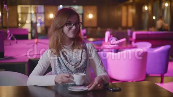 漂亮的带眼镜的年轻女士享受咖啡休息在餐厅放松喝咖啡看相机和表演视频的预览图