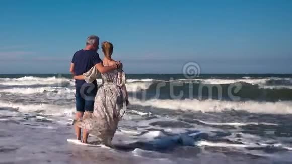 一对苗条的夫妇一个穿着牛仔短裤的男人和一个穿着漂亮长裙的女人站在沙滩上的大风中视频的预览图