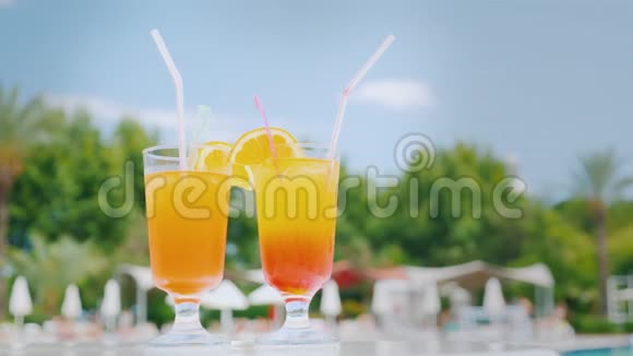 泳池边的沙滩桌上放着两杯带有吸管和橘子片的橙色鸡尾酒映衬着蓝天视频的预览图