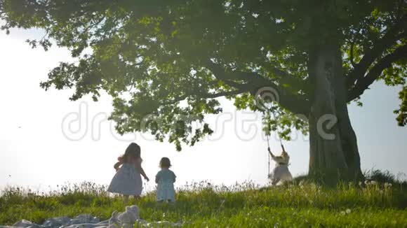 两个姐妹跑过干净的绿色田野走向母亲在一棵老橡树上荡秋千视频的预览图