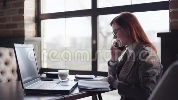 现代女教练建议一名在线学生使用手机一边坐在咖啡馆的桌子旁一边用电脑工作视频的预览图