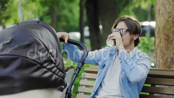 年轻的父亲正在荡着婴儿车喝着咖啡在公园里休闲视频的预览图