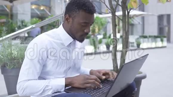 侧面观看自信的非裔美国人男子穿着白色衬衫在笔记本电脑上打字街道上的笔记本电脑上打字男性视频的预览图