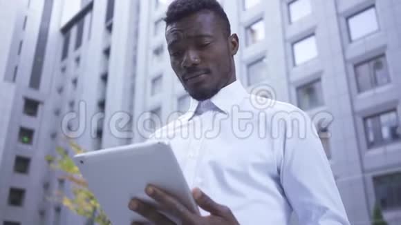 担心穿着白色衬衫的美国黑人男性在摩天大楼的背景上与平板电脑合作视频的预览图