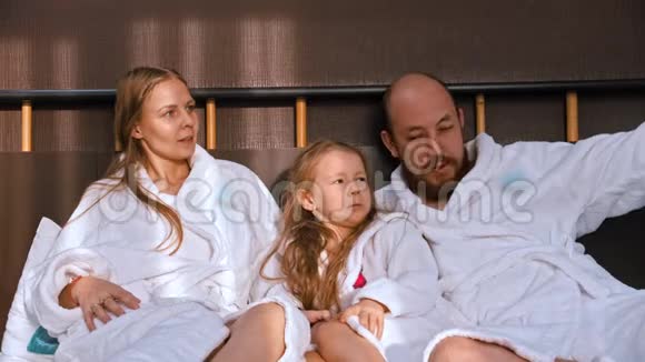 一个幸福的家庭穿着白色浴袍坐在床上互相交谈视频的预览图