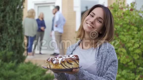 年轻迷人的女人带着蛋糕微笑着看着相机一群人在门廊上交谈视频的预览图