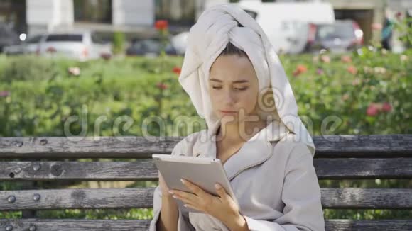 穿着浴袍头上戴着毛巾坐在公园里的长凳上检查的可爱微笑的年轻女子的画像视频的预览图