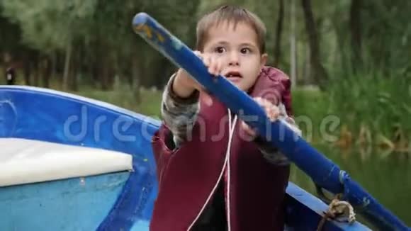 可爱的小孩子穿着救生衣坐在船上拿着木桨迷人的男孩试图驾驶和划船可爱的李视频的预览图