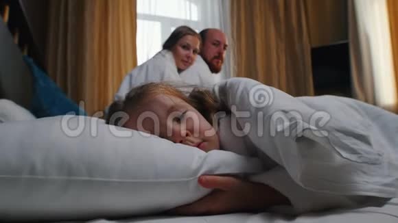 旅馆房间里的一家人一个睡在床上的小女孩她的父母看着她视频的预览图