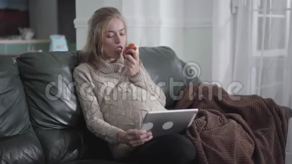 美丽的怀孕白人女孩吃苹果和看电视平板电脑年轻开朗的女人在沙发上休息视频的预览图