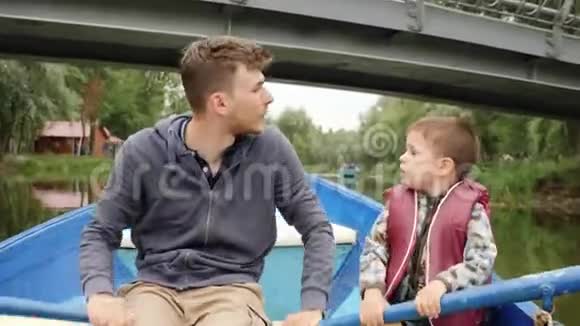 年轻快乐的父亲和他可爱的小儿子漂浮在人行桥下的木船上举起手来家庭幸福视频的预览图