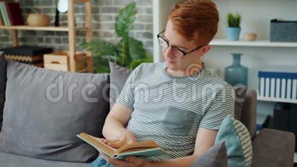 戴眼镜看书的年轻人坐在公寓的沙发上视频的预览图