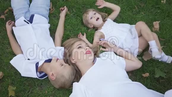 漂亮的金发女人和她的两个孩子躺在公园的绿草上小男孩小女孩漂亮女人视频的预览图
