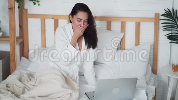 穿着浴袍的年轻女子坐在床上在家里用笔记本电脑工作打哈欠视频的预览图