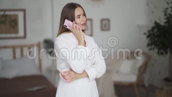 身穿白色浴袍的年轻女子喝茶在家打电话视频的预览图