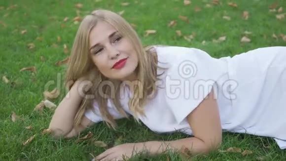 美丽的金发女人躺在草地上看着相机微笑着离开很漂亮的模特摆姿势户外休闲视频的预览图