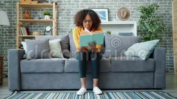 快乐的非裔美国学生阅读书籍和微笑在家里的沙发上放松视频的预览图