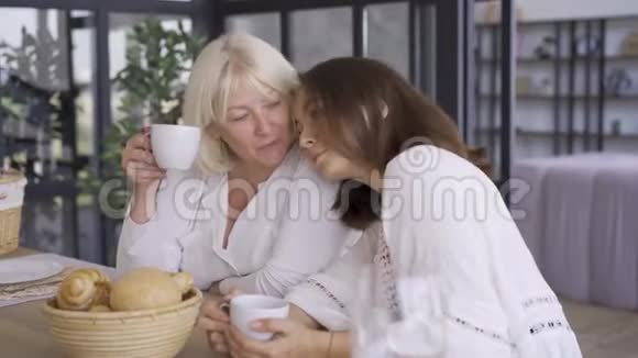 积极成熟的女人坐在厨房里女儿喝茶或咖啡把她的秘密和视频的预览图