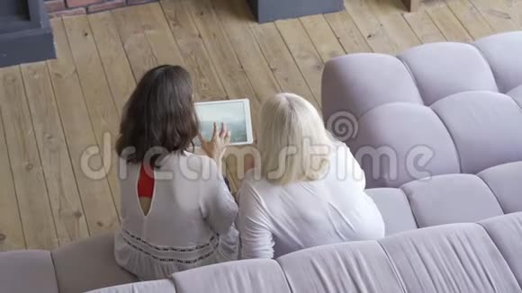 年轻女子和母亲坐在沙发上在平板电脑上展示她的旅行照片友好家庭视频的预览图
